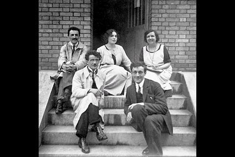 A black and white historical photo of scientists wearing lab coats sitting on the steps outside a building